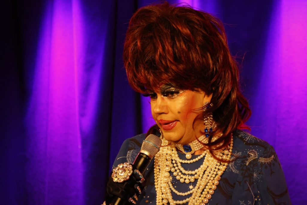 Drag queen med rødbrun paryk og perlekæder taler i en mikrofon foran lilla scenegardiner