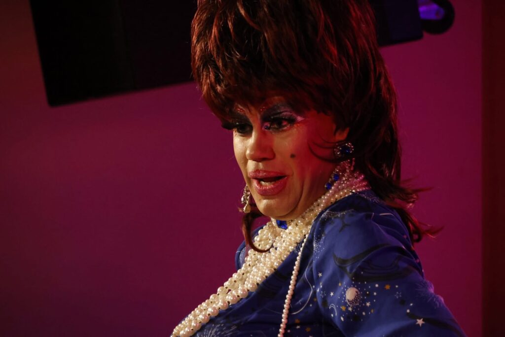 Drag queen med rødbrun paryk og perlekæder på scenen foran lyserød baggrund