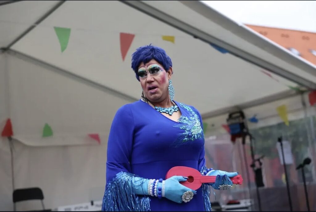 Drag queen i blå kjole og blå paryk med en rød ukulele foran et hvidt telt med flag