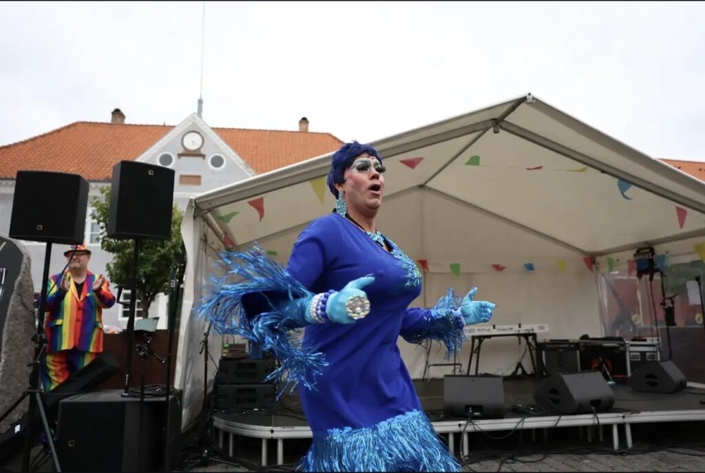 Drag queen i blå kjole og blå paryk danser på en lille scene foran et telt og en bygning med rødt tag